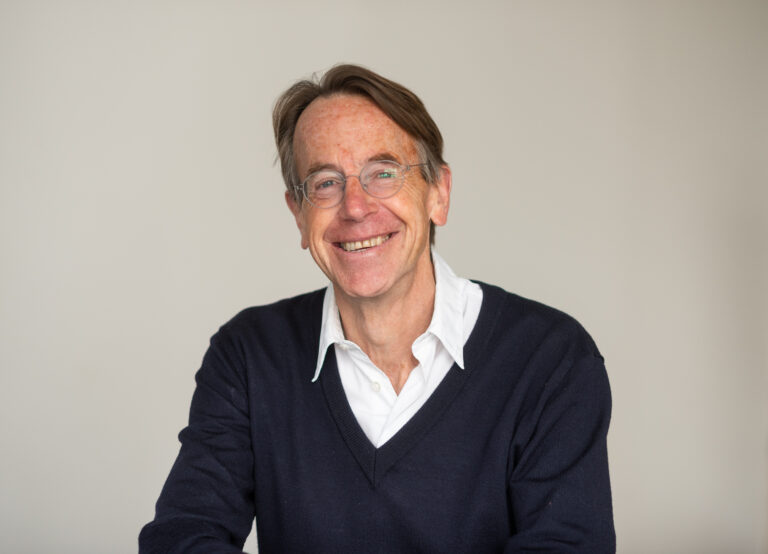 A white man with middle parted dark hair who wears glasses, a white shirt and dark sweater, is smiling at the camera