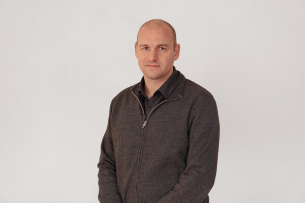 A white man wearing a grey jumper is standing in front of a neutral background. He is looking directly at the camera.