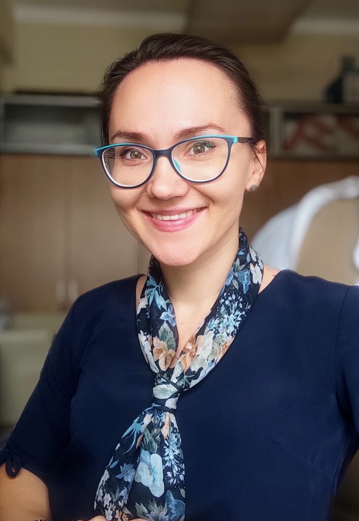 A white woman with dark hair who wears glasses is smiling at the camera