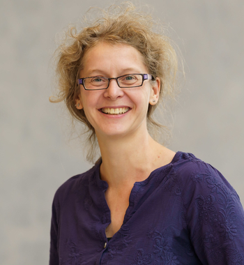 The picture of a white woman with curly blonde hair and glasses. She is smiling at the camera in front of a neutral background.