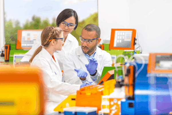 The picture of two women and a men in a laboratory. They all are wearing glasses, white medical gowns and light purple gloves.