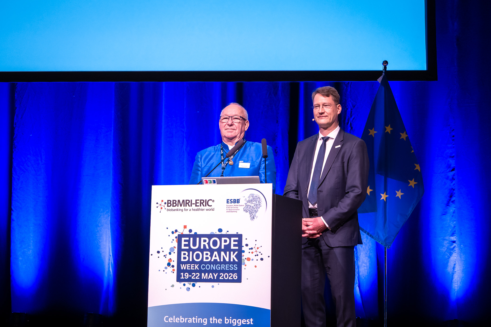 Two men are smiling at the audience from a stage